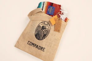 Burlap bag with 'Compadre' branding and a colorful serape on a beige background